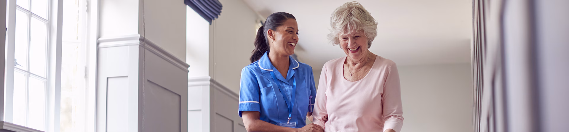 Senior Woman At Home Using Walking Stick Being Helped By Female Care Worker In Uniform
